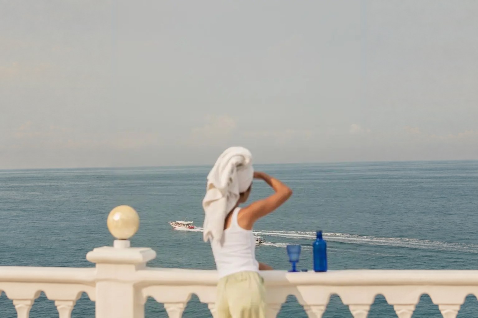 Una mujer con una toalla en el pelo mira el mar desde una balaustrada blanca, junto a una botella y una copa azules
